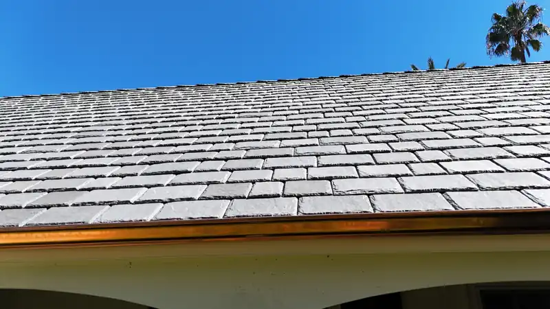 Gray slate-look composite tile roof edge with a copper gutter against a blue sky